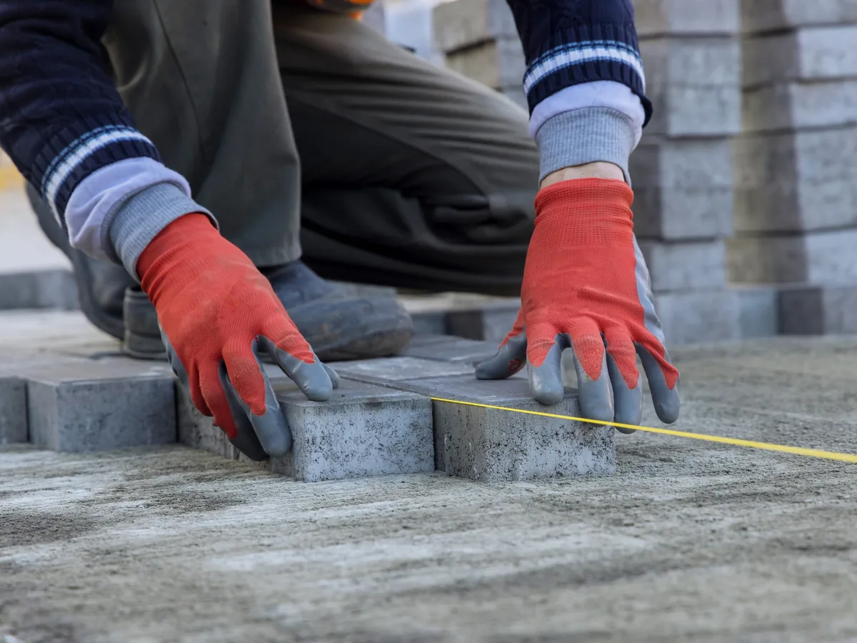 Gloved hands setting a grey paver to a string line with cut units stacked behind